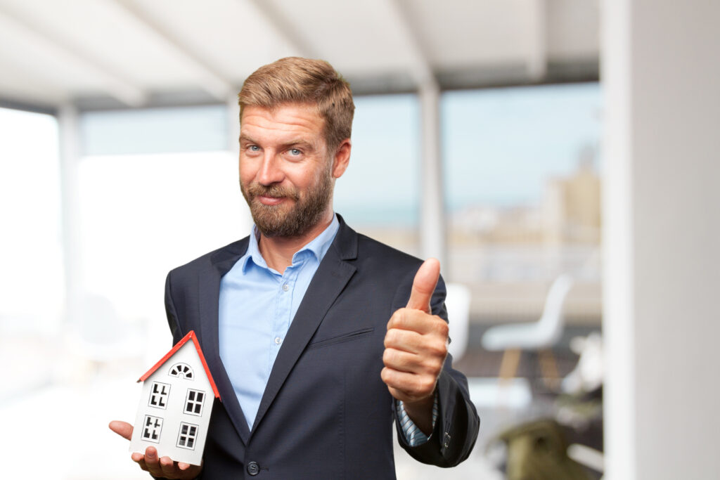 A smiling man with a beard, dressed in a dark blazer and blue button-down shirt, holds a small model house with a red roof in one hand while giving a thumbs-up with the other. The background is a bright, softly blurred office or living space.