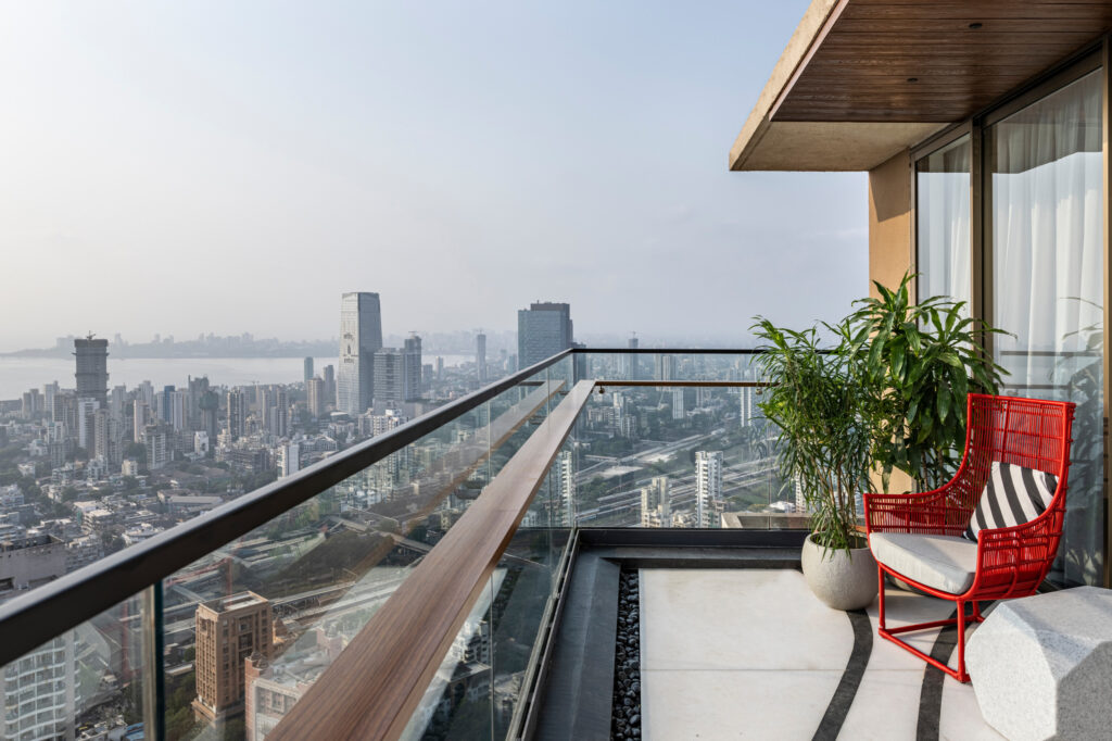Panoramic skyline view from a high-floor balcony of a luxury high-rise apartment, featuring a red designer chair and glass railings.