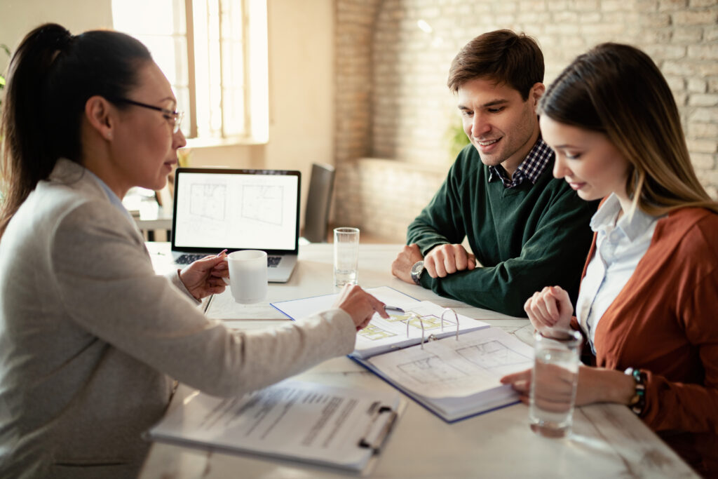A side-view of a female consultant in a grey blazer discussing architectural floor plans with a young couple. She is pointing to a specific detail in a binder while the couple looks on attentively. A laptop showing a digital version of the blueprint is open on the table between them.