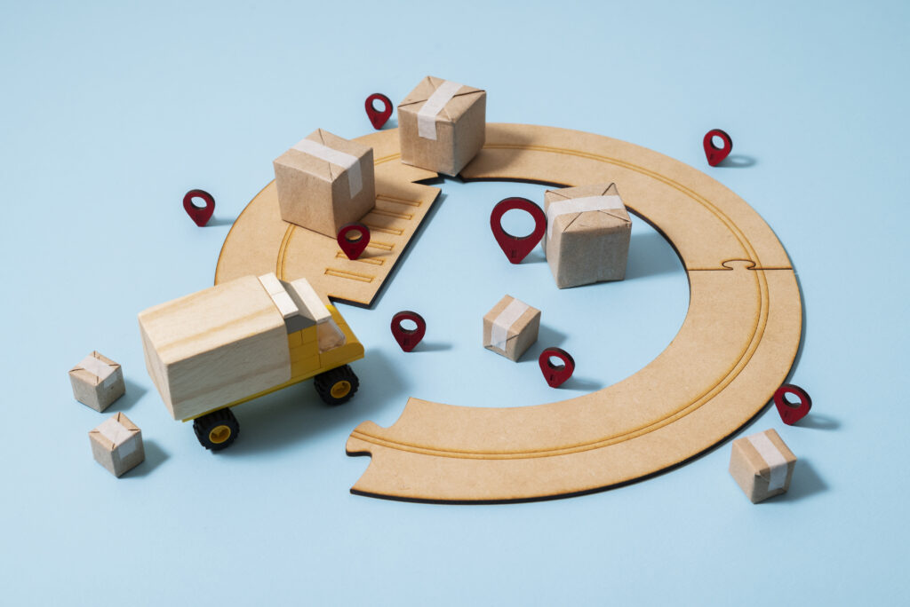 A stylized diorama on a blue background featuring a yellow toy truck driving on a wooden track, surrounded by cardboard boxes and seven red location pin markers.
